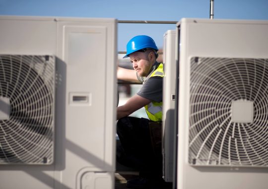 Technician in blue helmet inspecting an HVAC unit outdoors.